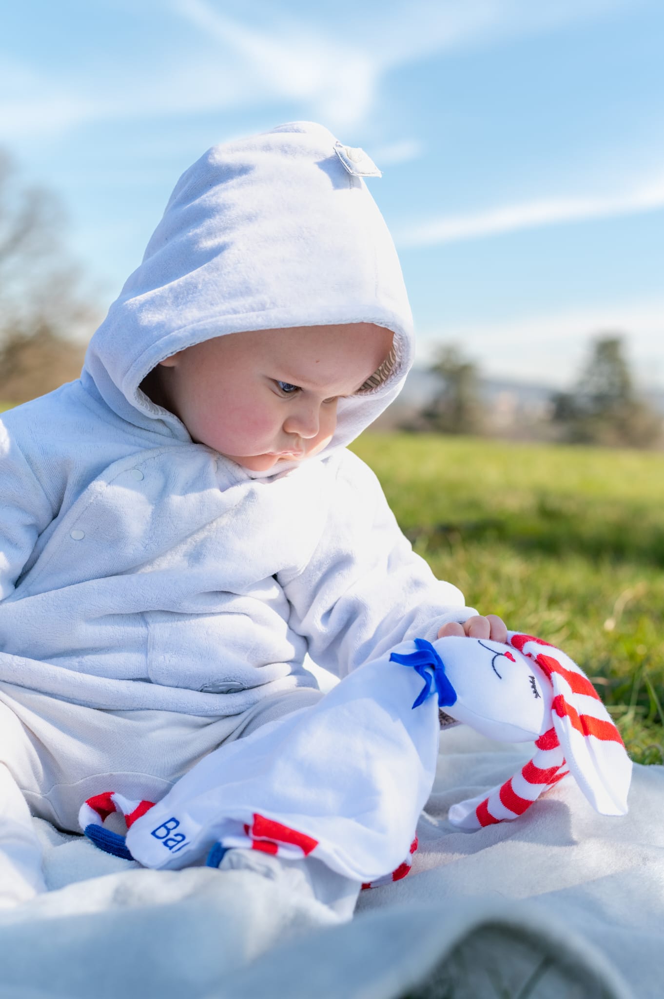 adorable bébé assis dans l'herbe avec capuche blanche qui regarde son doudou à rayures blanches et rouges