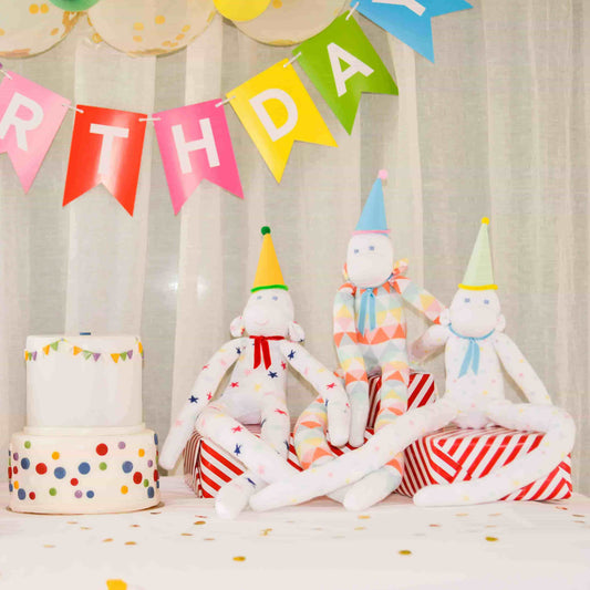 les doudous singes Marcel, Lucien et Léon sont à un anniversaire et portent des chapeaux de fête et sont assis à coté du gâteau