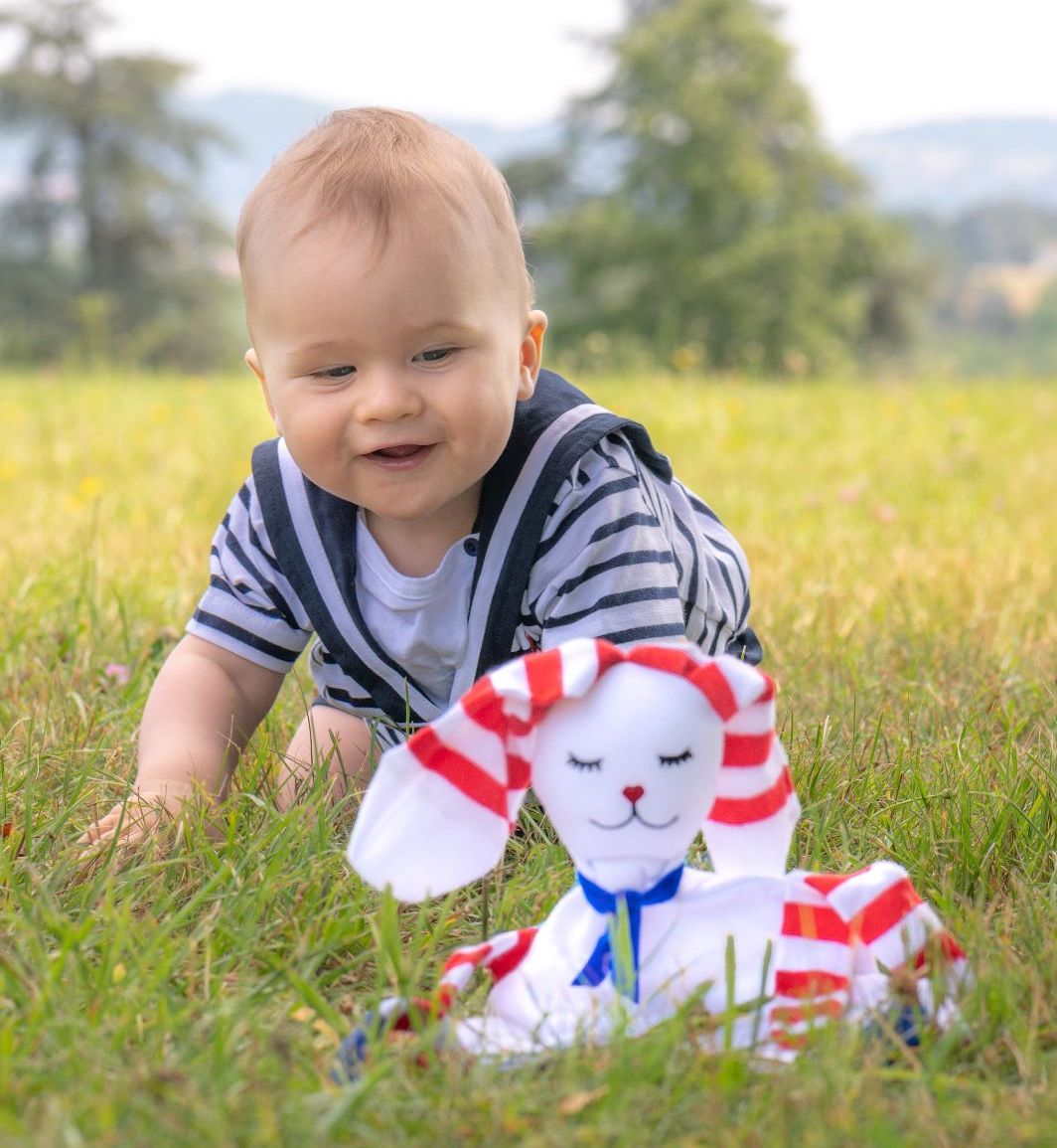 petit garçon blond de 6 mois qui joue avec un doudou lapin à rayures dans un jardin.jpg